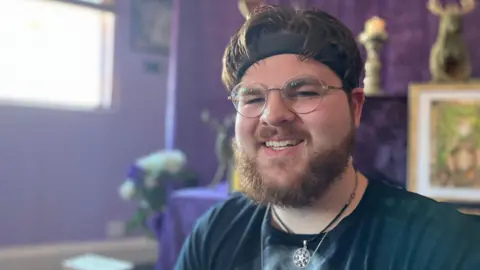 Thomas Clare smiles at the camera. He has a beard, wears glasses and a dark T-shirt with a pentagram necklace. The purple altar is behind him.