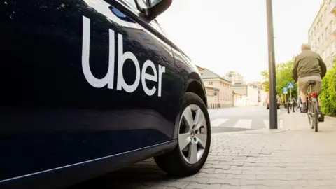 A black vehicle with the white Uber logo printed on its side is stopped, whilst a cyclist rides on the path next to it.
