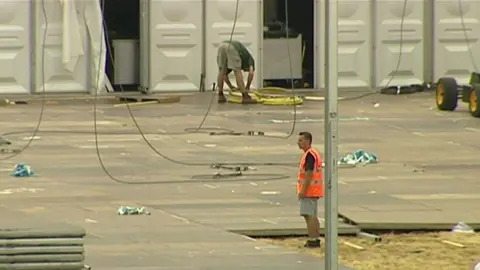 Two men working to set up the temporary mortuary. One of the men is bending down near what appears to be some yellow cables, while the other, wearing a bright orange safety vest, stands observing. The ground is scattered with cables, debris, and large panel-like tiles.