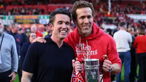 Rob McElhenney and Ryan Reynolds stand side by side holding a trophy on the Wrexham pitch and smiling. Ryan is wearing a red hoodie with Red Dragons written in white on it and Rob wears a black short-sleeved polo shirt