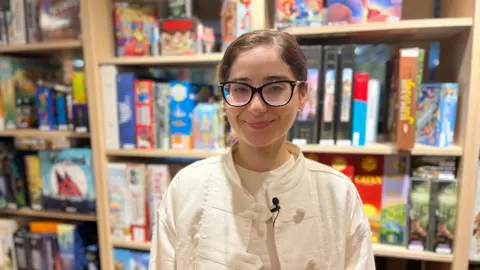Melika Latif stands in front of rows of colourful board games. She wears a white military style jacket with a white t-shirt underneath. She has brown hair tied back and wears glasses.