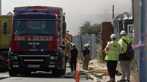 A red appliance with Fire & Rescue written across the front and blue lights flashing is parked on a road with hoses running across the street. Smoke is rising from behind a nearby fence. Several firefighters in uniform are working around the truck, and a number of other people in hi-viz jackets and white helmets are looking on.