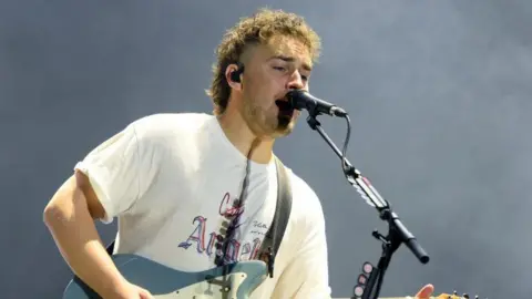 Getty Images Sam Fender wearing a white t-shirt singing into a black microphone with a black ear piece in his right ear.