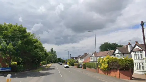 Google Alcester Road, a single lane highway with trees on either side and white houses on the right hand side.