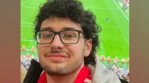 A young man at a football stadium. He is wearing a red scarf. 