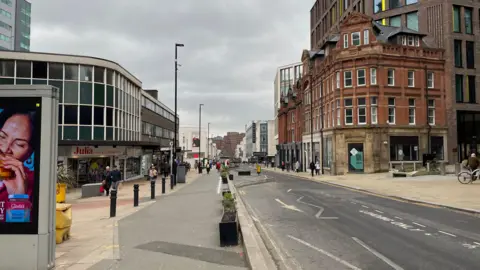 A high street through a city centre has two sides of a road with a large concrete boundary and planters down the middle. There are shops on either side of the road