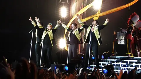Getty Images Take That stand at the front of a stage during a live performance. The four members have their arms raised up into the air. The arena seems largely dark other than the spotlight shining on the band. 