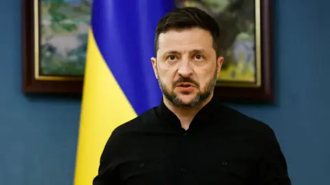 Reuters Zelensky speaking during press briefing following calls with Trump. behind him is a blue wall, a picture frame and a Ukrainian flag