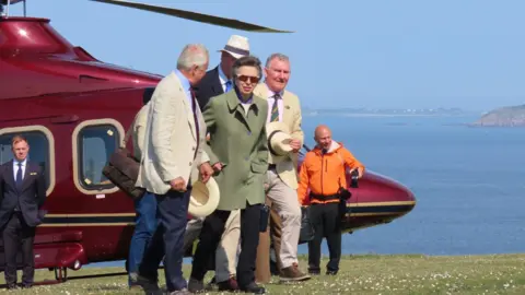 The Princess Royal arriving in Sark in the Channel Islands. She is surrounded by several others and is walking away from a burgundy helicopter wearing a khaki green coat and black trousers.