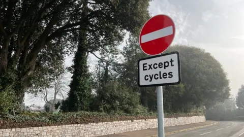 A red and white no entry sign that says 'Except cycles' underneath at the part of Dryden Road which has barriers stopping motorised vehicles from entering.