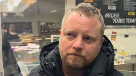 Elliot Deady/BBC Jamie Towells looks away from the camera to the left as he stands in his butchers. He has short grey hair and wears a black rain coat with a large hood.