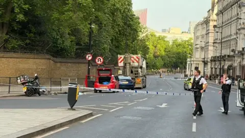Two policemen are seen in front of a cordon on Grosvenor Place. A car boot in seen open, while debris is seen in the road 