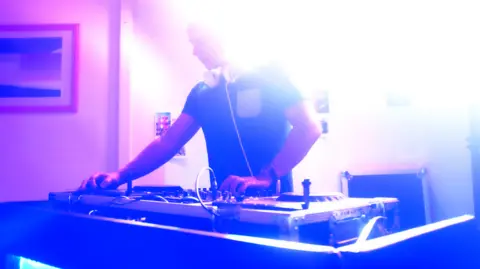 BBC A picture of a DJ. He is wearing headphones and is standing over DJ decks.