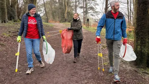 Three people walking on a path in an area full of trees. A woman in the middle is holding a large plastic bag, wearing dark trousers and green jacket. A man to the right is wearing a blue jacket, light trainers and a red top and is holding a plastic bag. A woman to the left is also holding plastic bag, wearing blue jeans, red top, blue jacket and blue hat. 