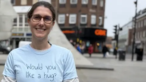 A smiling woman wearing glases and a blue t-shirt with the words "Would you like a hug?" stands in the street.