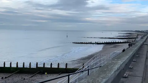 View of the beach from the promenade with water lapping up by the side of groynes. 