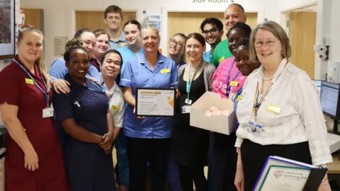 A group of medical staff at RUH Bath. They are all pictured standing close together in a group, smiling at the camera. Many of them are wearing scrubs, but one woman is wearing a pink Organ Donation t-shirt. One woman is holding a framed certificate and another is holding a box of pink cakes. 