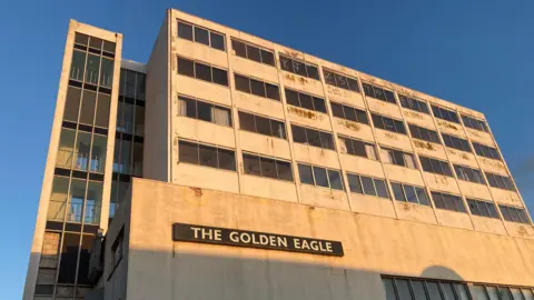 LDRS A tall stone building with smashed windows. A sign on the front reads' The Golden Eagle'. The sun is setting on the side of the building. 
