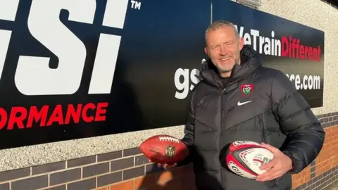 A man in a black jacket carries an American football and a rugby ball