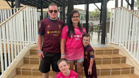 Jamie Warden stands on steps at a hotel with his wife and their two young boys. They are all wearing Ipswich Town football shirts. Jamie wears sunglasses and has short dark hair and shorts. His wife has long dark hair and is wearing shorts. The two boys both wears glasses, have short dark hair and are also wearing shorts. 