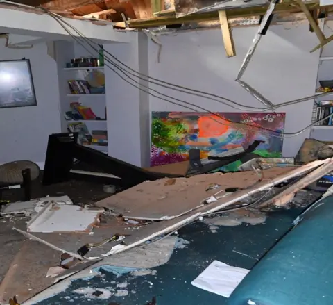 PA The roof has collapsed on what appears to be a brown sofa in a living room. A colourful painting can be seen in the background amidst a mess of debris. 