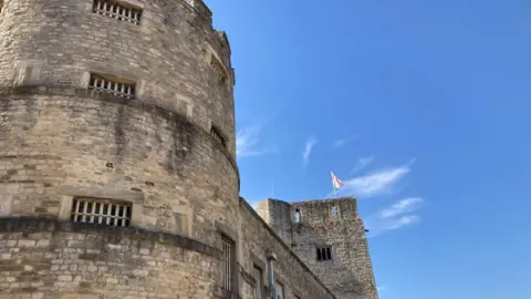 An old castle building is in the foreground with a blue sky behind. The cross of St. George is flying from the top of the tower.