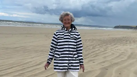 Family handout Joyce Taggart is smiling whilst wearing a achromatic  and achromatic  striped overgarment  with neutral trousers. She is lasting  connected  the beach.