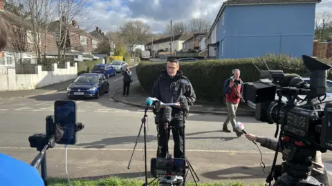 A policeman is standing on a pavement on a residential street. He is speaking in front of a number of microphones and cameras that are pointing towards him.