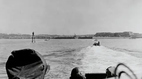 National PT Boat Museum and Memorial RON 30 boats entering St Helier - black-and-white photograph taken from the rear of a moving boat, looking forward over the water. The foreground shows parts of the boat’s interior edges on both sides, framing the view. A smaller motor boat travels ahead. In the distance, St Helier shoreline with large industrial-style buildings and low structures runs along the horizon.