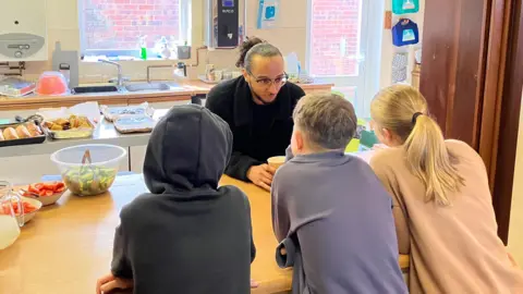 Jon Wright/BBC Isiah Jordan is one side of a kitchen hatch - there are three young teenagers the other side, with their backs turned to the camera. There is salad and bread on the counter. Isiah has a black coat on and black-rimmed glasses. 