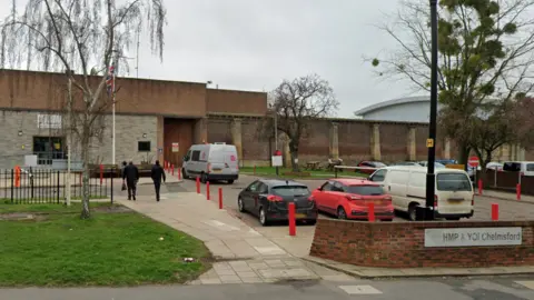 Google HMP Chelmsford seen from the street. It has large brick walls and has cars parked outside. There is a silver sign that reads "HMP & YOI Chelmsford".