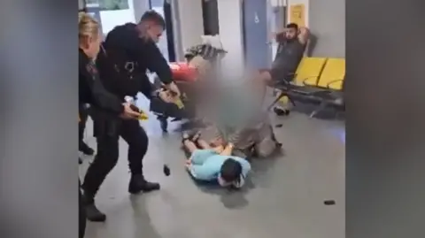 mobile screengrab of two officers pointing Taser at man lying Tasered on ground, with female relative crouching over him and another man sitting behind with his hands behind his head