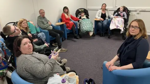 A group of mothers and fathers sit in a circle in blue bucket chairs. A few children are sitting between them, two are in prams. The parents are looking at the camera but not smiling.