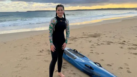 Young girl standing on beach with the waves and skyline behind her and her rescue board by her side 