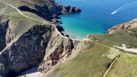 La Coupée in Sark on a sunny day. It is a narrow strip of land that goes over a cliff. The sea is next to the area and a speedboat is heading towards the island.