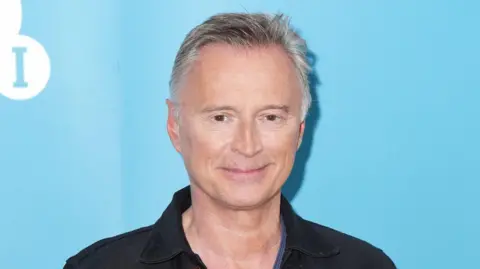 Robert Carlyle has short white hair and smiles. He wears a black top and stands in front of a blue background 