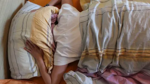 Getty Images A person in a bed covers their head with a pillow as if trying to avoid a loud noise. The bed linen is white and grey with yellow stripes.