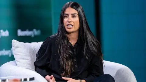 Bloomberg via Getty Images Asha Sharma, wearing a black top and trousers, sits on stage speaking to an interviewer out of shot at a Bloomberg Tech conference.