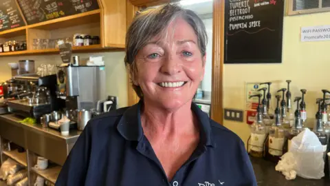 A woman with medium length grey hair looks to the camera as she wears a navy polo shirt. She has blue glasses resting on her head. Behind her right shoulder is a silver coffee machine, below it are silver shelves with white mugs. Above the machine are blackboards with different items written on it. 
