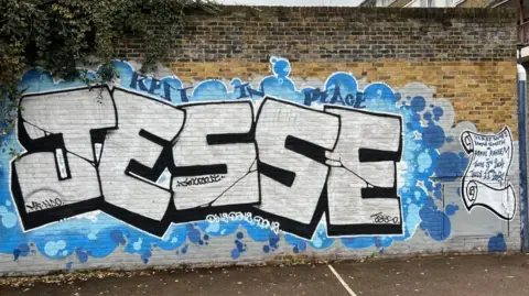 A blue and white spray-painted mural on the side of the brick wall of a garage block, which says 'Jesse'