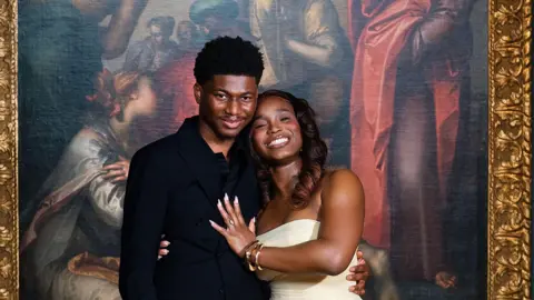 Emmanuel Dosumu/adidolvisuals Emmanuel and Elizabeth stand holding each other and smiling in front of the painting where they got engaged.