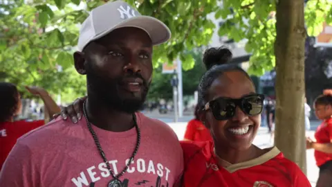 Jack Valpy/BBC A man in a pink shirt and a gray New York Yanke's hat. He stands next to a woman with bound black hair and black sunglasses. She wears a red -gold white town FC shirt.