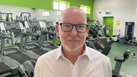 Man with bald head, glasses and white beard. He is standing in front of gym equipment.