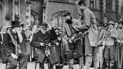 Topical Press Agency/Getty Images A black and white archive photo of Winston Churchill visiting Queen's' University, Belfast in September 1926.  He is seated on a small stage surrounded by a crowd of onlookers.  He is wearing a dark overcoat with a large fur collar.  Churchill is being presented with a hat and a clay pipe by students during rag week. One of the students is wearing a top hat and a long, fake beard. 