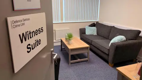 A witness suite at the DSCU headquarters in Bulford, where victims and witnesses are interviewed. It is a relaxed, comfy room with a black sofa, blue fluffy cushions, a coffee table and a potted plant.