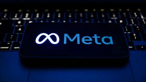 Getty Images Smartphone displaying Meta's logo in blue, glowing against a black background. The phone is lying on a lit-up laptop keyboard. 