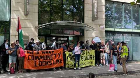 A group of people with banners referring to community stand outside a hotel entrance.