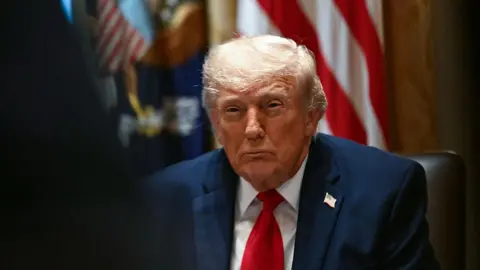 Donald Trump looks straight ahead with his eye narrowed. He is wearing a navy suit, white shirt and red tie with an American flag pin on his lapel. In the background is an American flag.