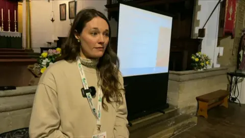 Emily Fedorowycz in a beige jumper stood in a church. She has long brown hair and is wearing a lanyard.