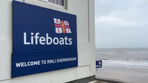 Andrew Turner/BBC The RNLI sign bears the charity's flag, which features a crown and anchor within a red cross of St George, with blue fimbriation and a white background featuring the initials RNLI. The words: "Lifeboats" and "Welcome to RNLI Sheringham" feature below the flag. The sign is mounted on the eastern wall of the lifeboat station, a concrete block building, that is overlooking the beach, with the beach and breakers in the backdrop.